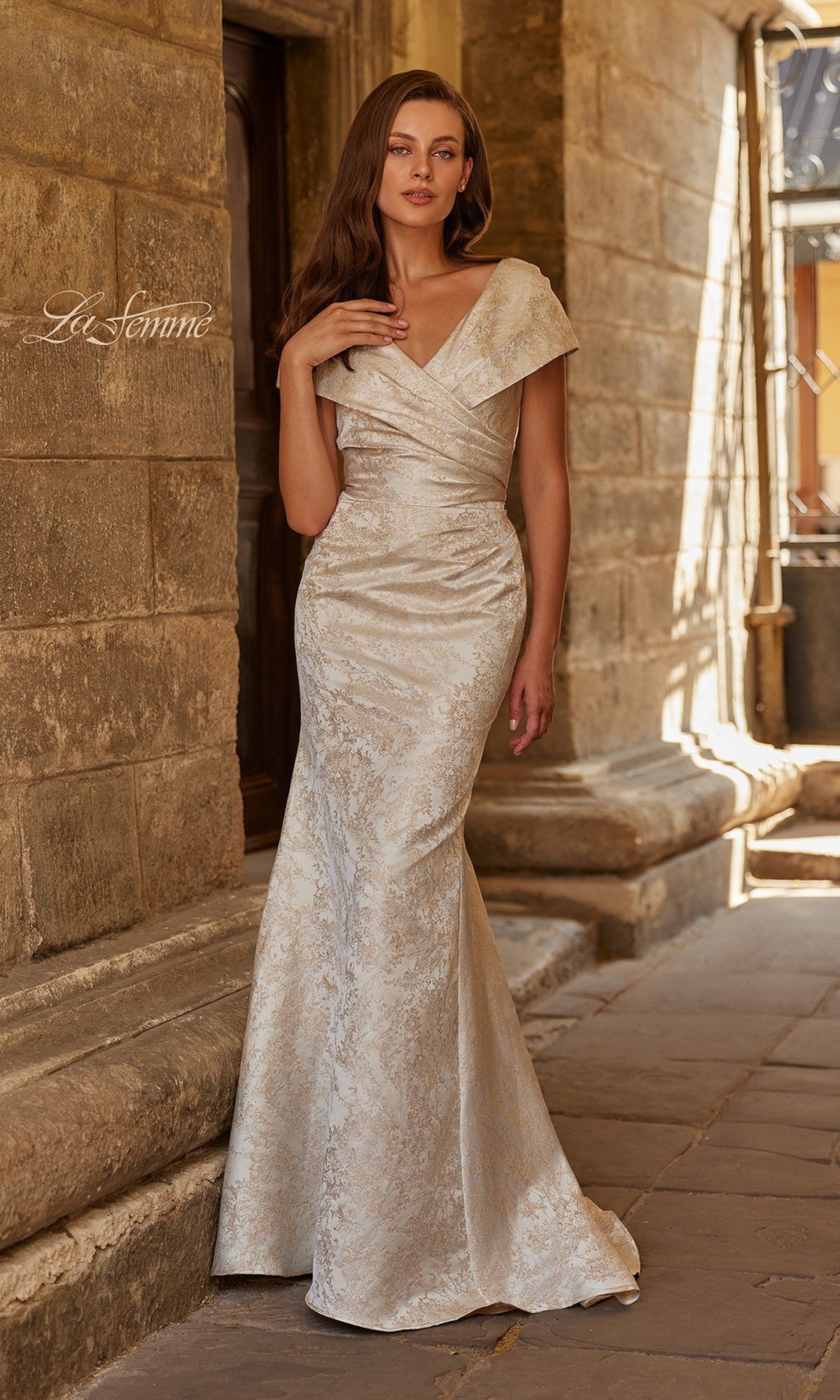 A woman poses outdoors beside a stone building, wearing the La Femme 33428 long formal dress in nude—a fitted, floor-length jacquard gown with a subtle floral pattern and an off-the-shoulder design.